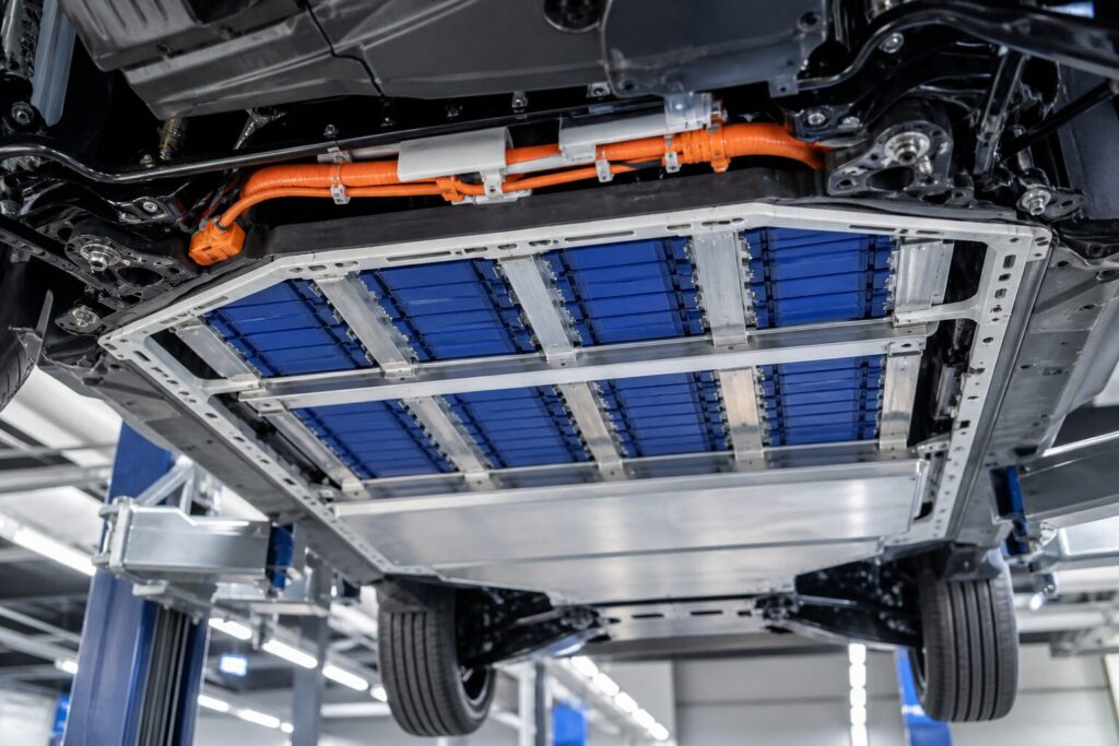 Pack de bateria de carro elétrico exposto sob um veículo elevado, com células azuladas e cabos de alta tensão, em oficina iluminada por luz técnica fria.
